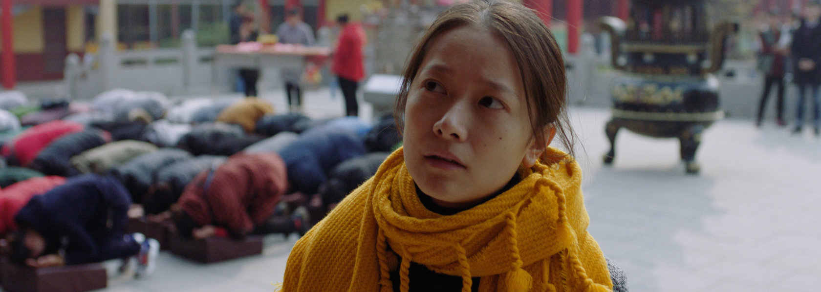 A young woman looking with eyes to her right with people kneeing down in prayers on background - Lost Lotus - THIS Buddhist Film Festival