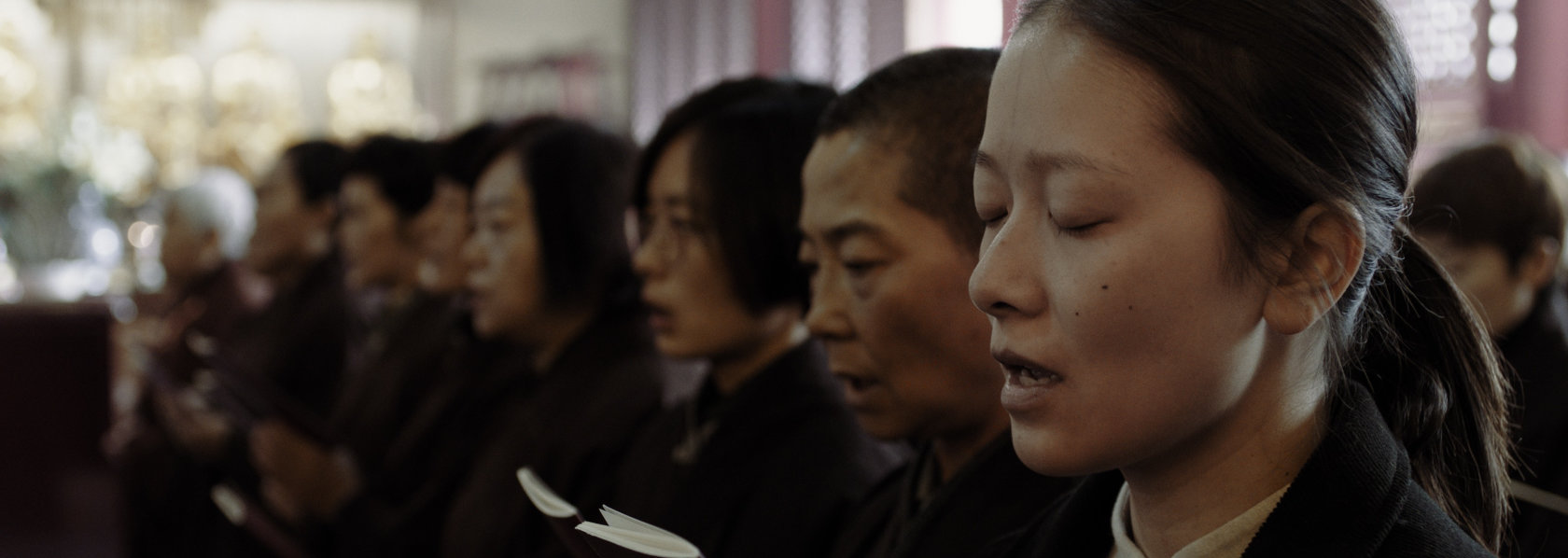 A young woman hold a chanting book with palms together with other laypeople - Lost Lotus - THIS Buddhist Film Festival